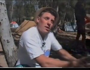 A young man in T-shirt and sun hat in a sunny camp site looking up at the camera.